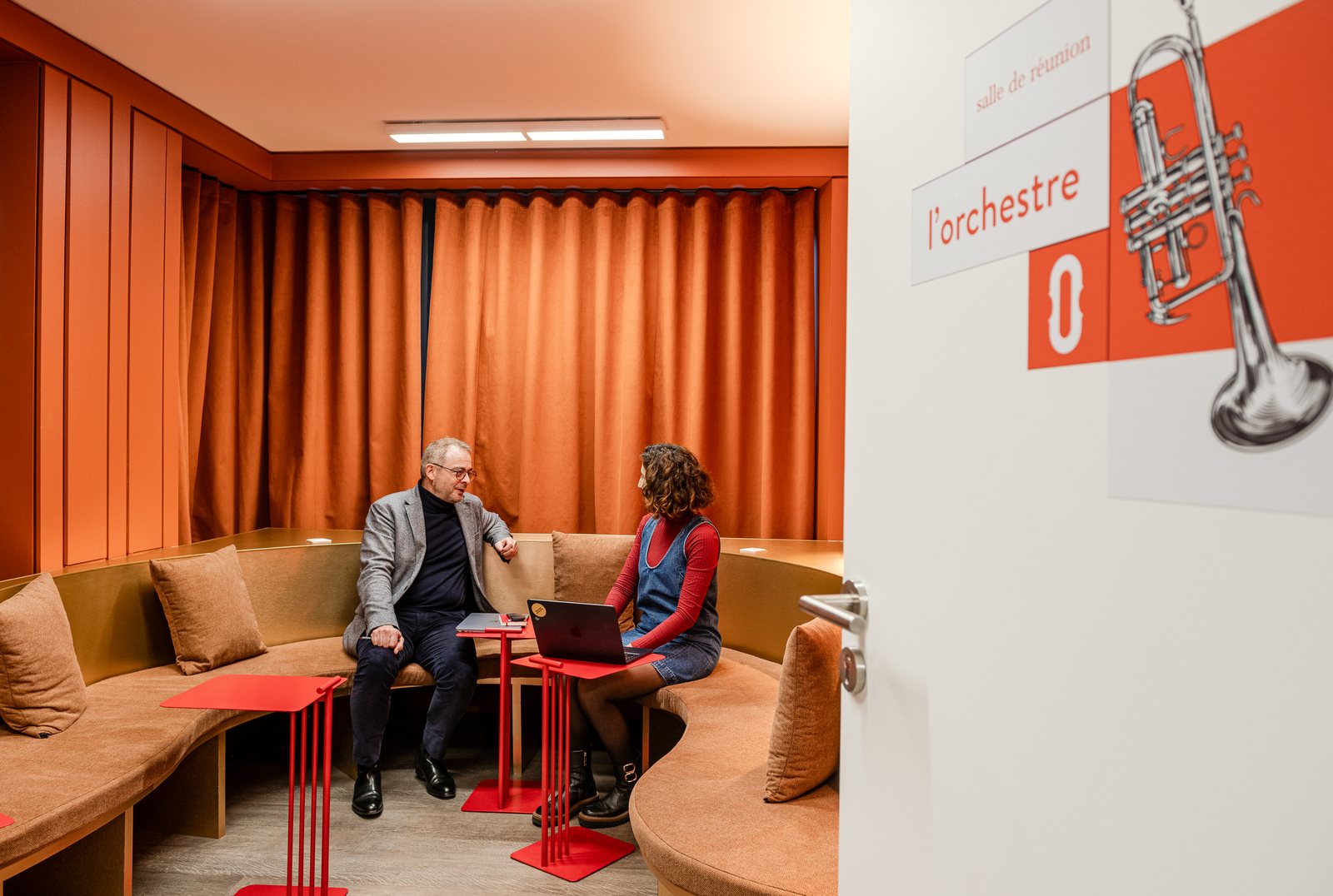 Salle de réunion L'Orchestre : banquette circulaire, rideaux de velours terracotta, mange-debout rouges et signalétique trompette. Deux personnes en réunion.